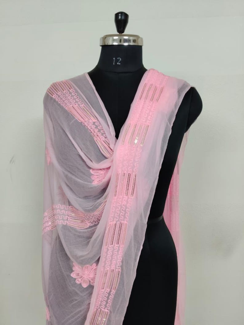 May include: A sheer, light pink scarf with intricate embroidered details and sequin accents. The scarf is draped over a black mannequin, showcasing its delicate floral and geometric patterns. The fabric appears lightweight and flowing.