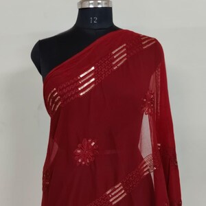 May include: A burgundy-colored sheer scarf draped over a mannequin. The scarf features gold-colored sequin embellishments in a linear and floral pattern. The fabric is lightweight and appears to be flowing.