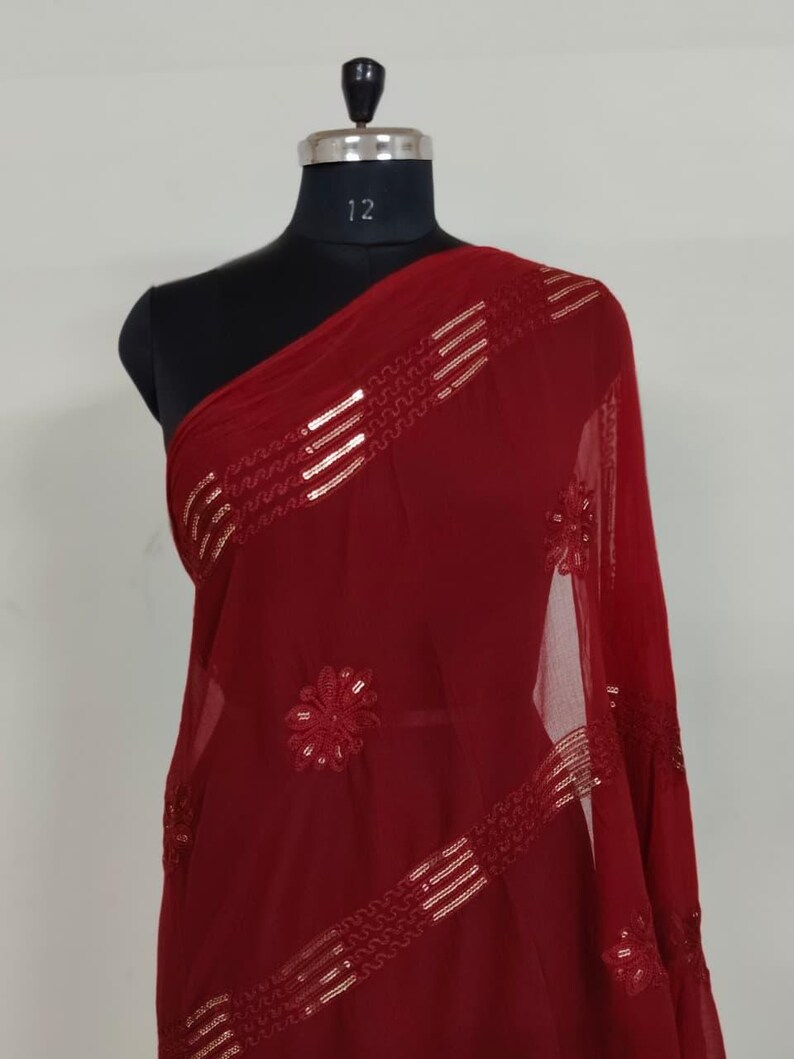 May include: A burgundy-colored sheer scarf draped over a mannequin. The scarf features gold-colored sequin embellishments in a linear and floral pattern. The fabric is lightweight and appears to be flowing.