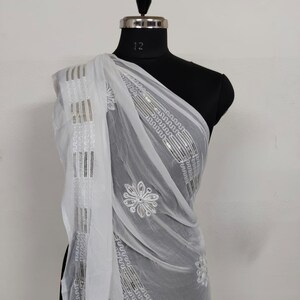 May include: A sheer white scarf draped over a mannequin. The scarf features embroidered floral and geometric patterns with silver accents. The fabric is lightweight and flowing, suggesting a delicate texture. The scarf is a fashion accessory.