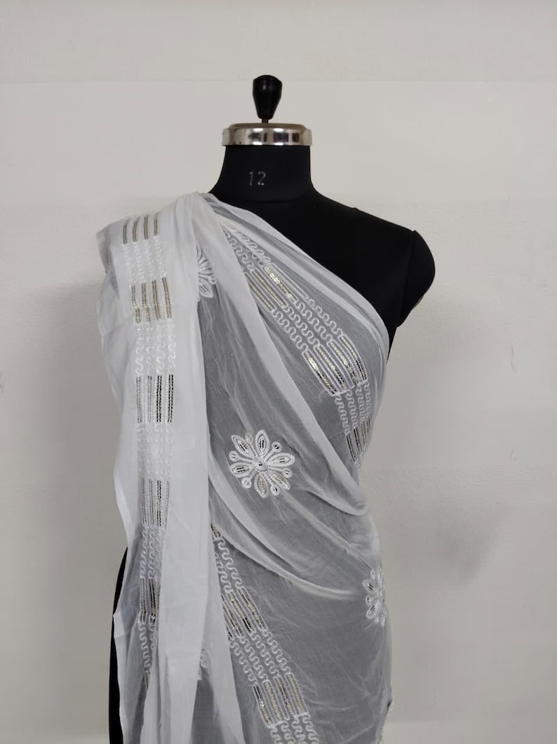 May include: A sheer white scarf draped over a mannequin. The scarf features embroidered floral and geometric patterns with silver accents. The fabric is lightweight and flowing, suggesting a delicate texture. The scarf is a fashion accessory.