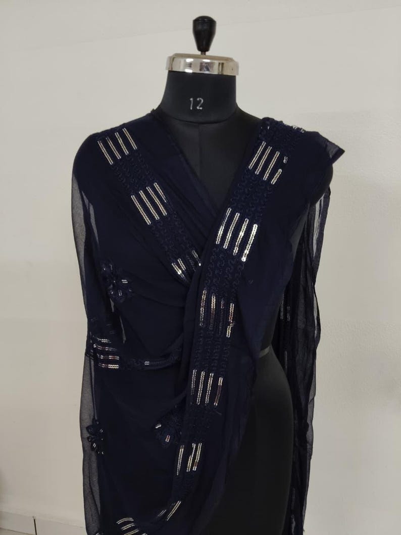 May include: A navy blue sheer scarf with silver sequin embellishments. The scarf is draped over a mannequin, showcasing the rectangular and floral patterns of the sequins. The fabric appears lightweight and flowing, suitable for formal wear.