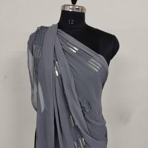 May include: A draped, gray chiffon scarf with silver sequin embellishments. The scarf is draped over a black mannequin, showcasing the fabric's flow and the intricate detailing of the sequins. The design features horizontal and vertical sequin patterns.
