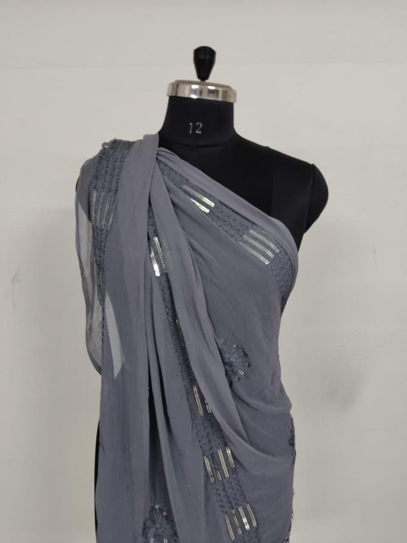 May include: A draped, gray chiffon scarf with silver sequin embellishments. The scarf is draped over a black mannequin, showcasing the fabric's flow and the intricate detailing of the sequins. The design features horizontal and vertical sequin patterns.