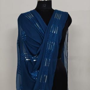 May include: A draped, teal-colored scarf with intricate sequin detailing. The scarf is displayed on a black mannequin, showcasing its flowing fabric and embellished design. The sequins are arranged in a linear pattern along the edges.