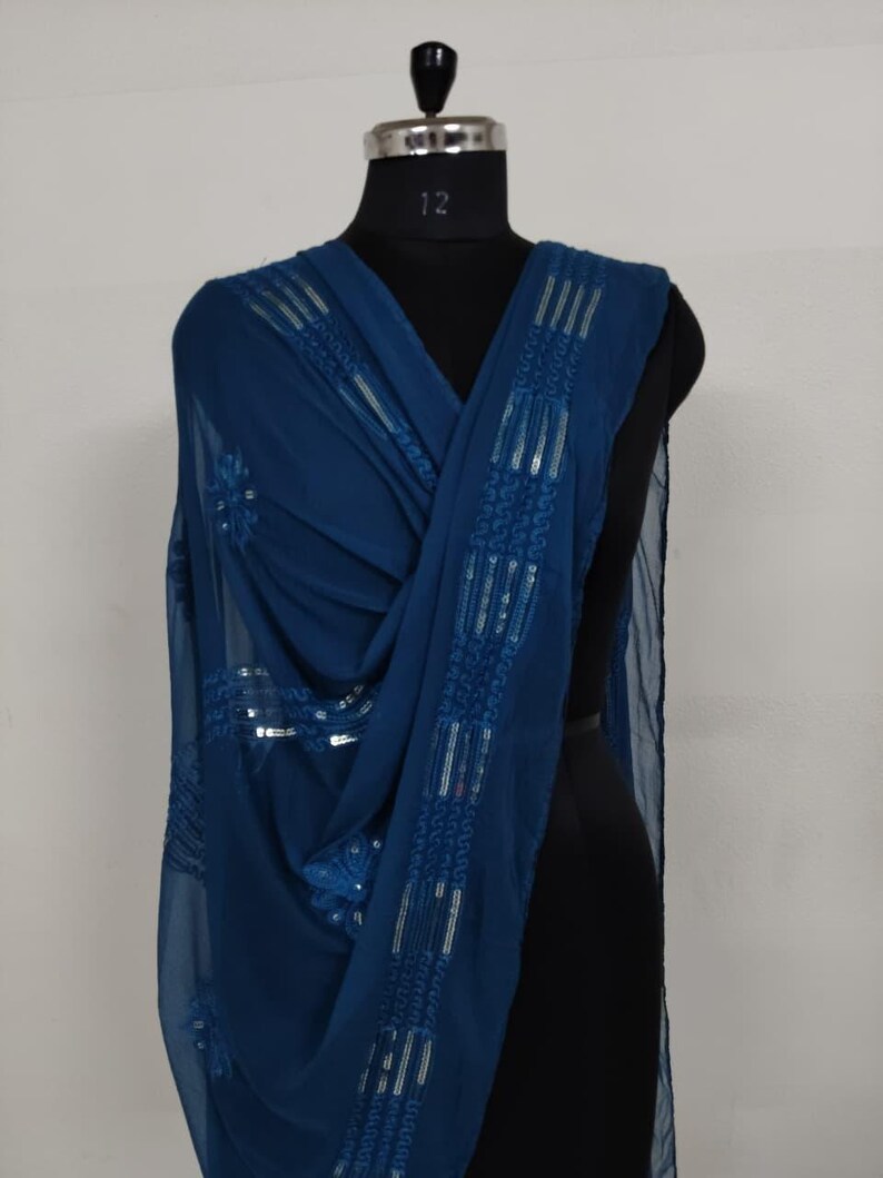 May include: A draped, teal-colored scarf with intricate sequin detailing. The scarf is displayed on a black mannequin, showcasing its flowing fabric and embellished design. The sequins are arranged in a linear pattern along the edges.
