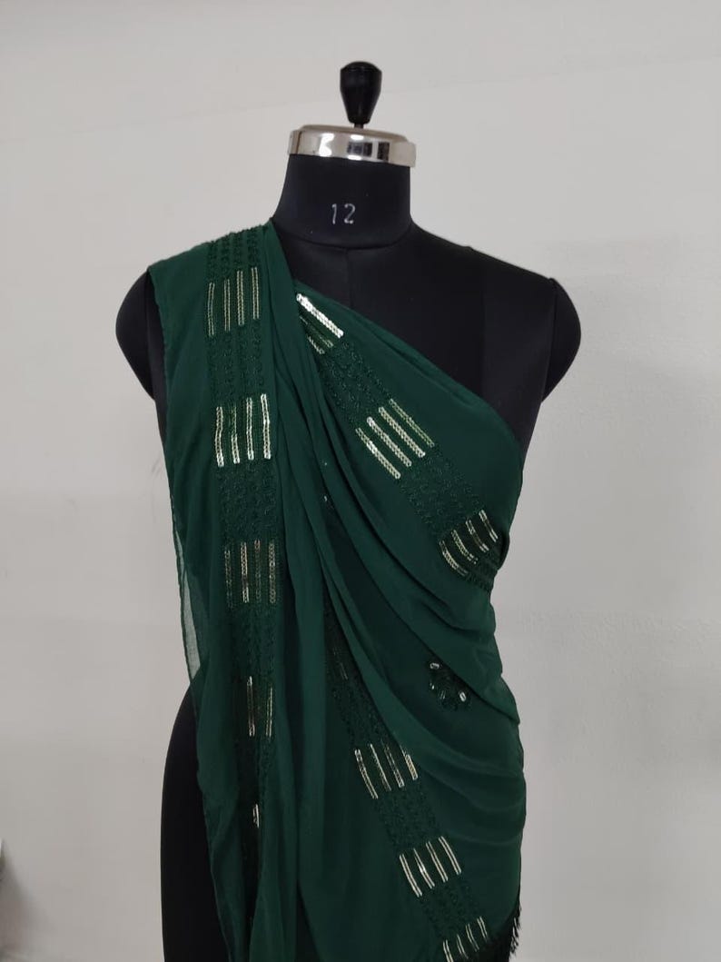 May include: A dark green, one-shoulder garment draped over a black mannequin. The fabric is embellished with vertical rows of silver sequins. The garment is likely a shawl or a piece of formal wear.