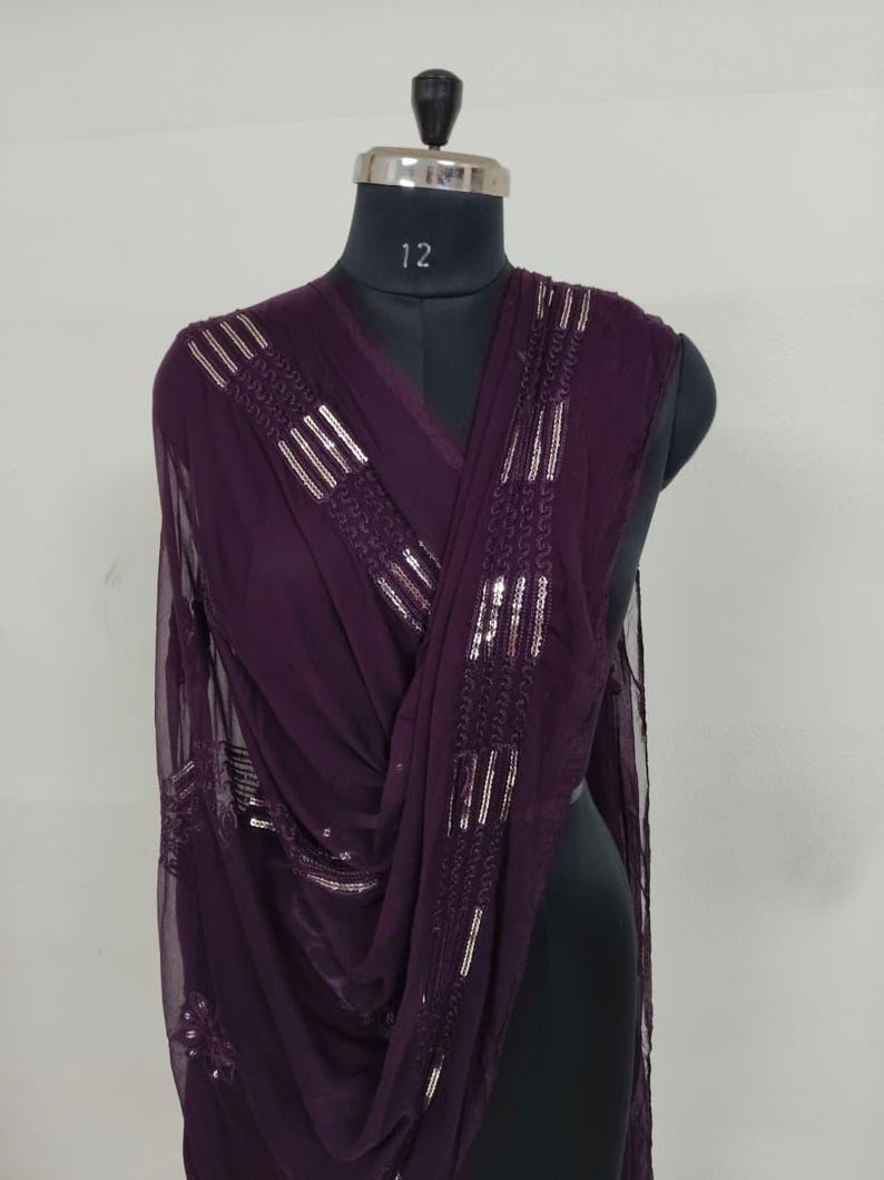 May include: A draped, plum-colored scarf with silver sequin embellishments. The scarf is displayed on a black mannequin, showcasing its flowing fabric and decorative details. The sequins are arranged in vertical and horizontal patterns.