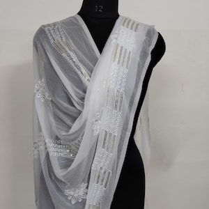 May include: A sheer white scarf with intricate embroidery and sequin detailing. The scarf is draped over a black mannequin, showcasing the delicate floral and geometric patterns. The design includes vertical stripes and floral motifs.