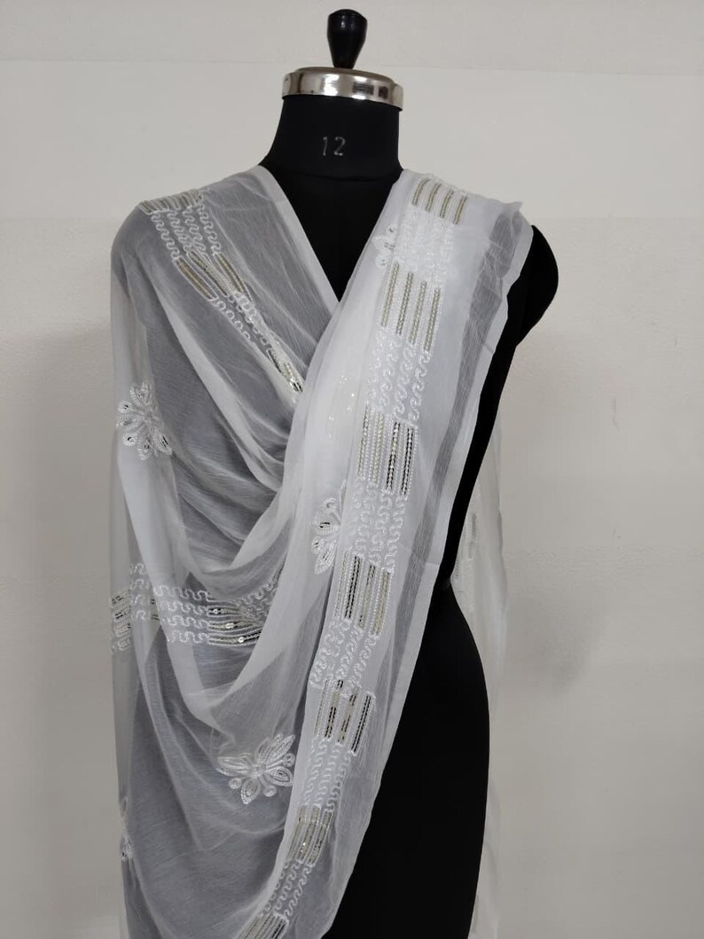 May include: A sheer white scarf with intricate embroidery and sequin detailing. The scarf is draped over a black mannequin, showcasing the delicate floral and geometric patterns. The design includes vertical stripes and floral motifs.