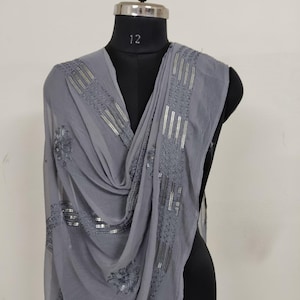 May include: A gray scarf with intricate silver sequin and embroidered detailing. The scarf is draped over a black mannequin, showcasing its flowing fabric and decorative embellishments. The design includes floral and linear patterns.