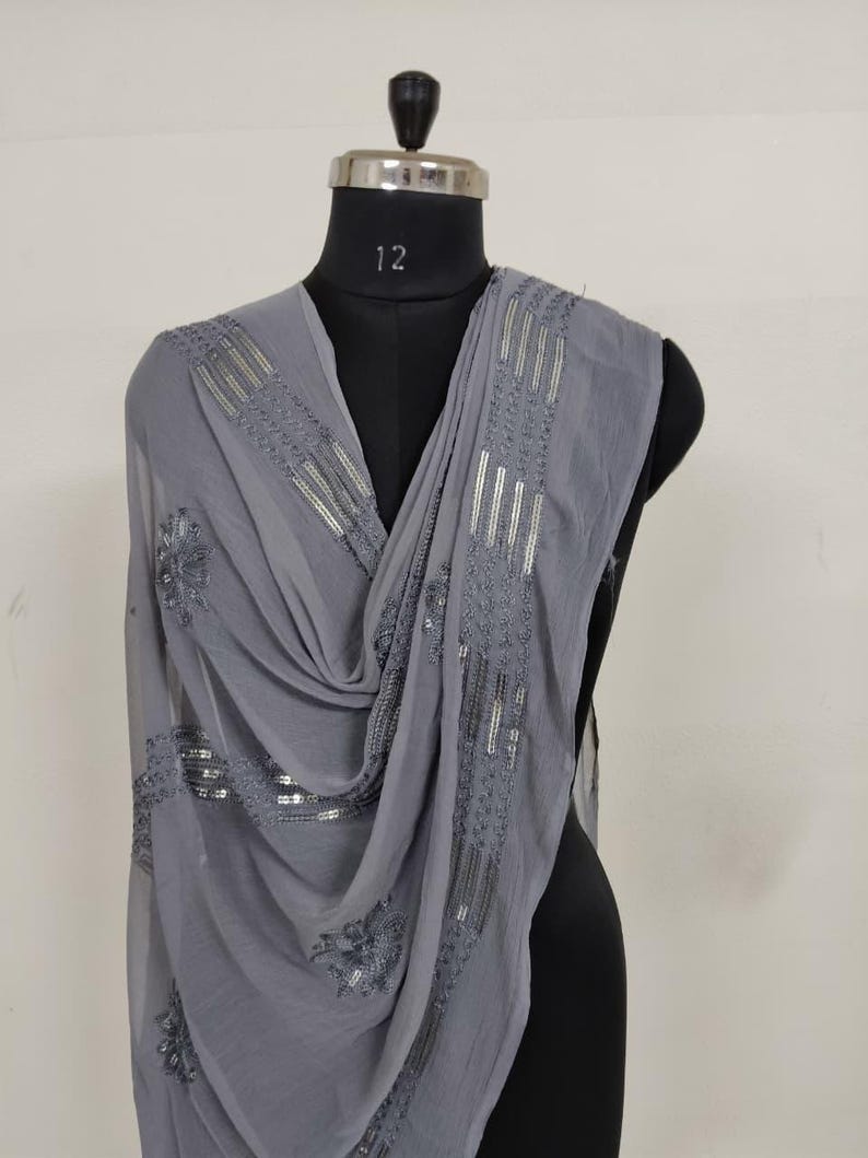 May include: A gray scarf with intricate silver sequin and embroidered detailing. The scarf is draped over a black mannequin, showcasing its flowing fabric and decorative embellishments. The design includes floral and linear patterns.
