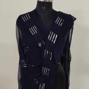 May include: A navy blue sheer scarf with silver sequin embellishments. The scarf is draped over a mannequin, showcasing the rectangular sequin patterns. The fabric appears lightweight and flowing, suitable for formal wear or adding a touch of elegance.
