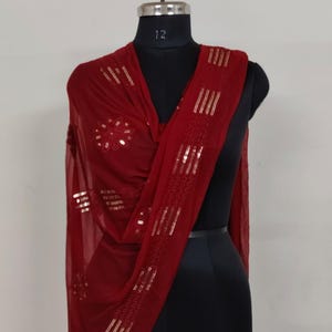 May include: A burgundy scarf with gold sequin embellishments is draped over a black mannequin. The scarf features a floral design and vertical stripes of sequins. The fabric appears lightweight and flowing, suggesting a delicate texture.
