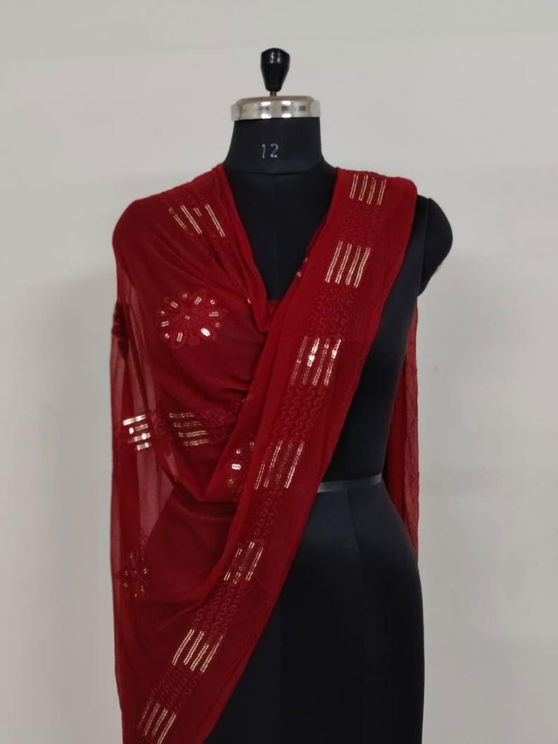 May include: A burgundy scarf with gold sequin embellishments is draped over a black mannequin. The scarf features a floral design and vertical stripes of sequins. The fabric appears lightweight and flowing, suggesting a delicate texture.