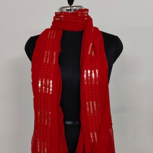 May include: A vibrant red scarf with gold sequin accents, draped on a black mannequin. The scarf is made of a lightweight fabric and features vertical rows of shimmering sequins, adding a touch of elegance to the design.