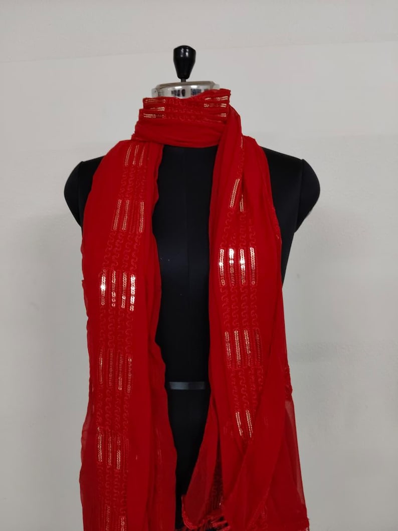May include: A vibrant red scarf with gold sequin accents, draped on a black mannequin. The scarf is made of a lightweight fabric and features vertical rows of shimmering sequins, adding a touch of elegance to the design.