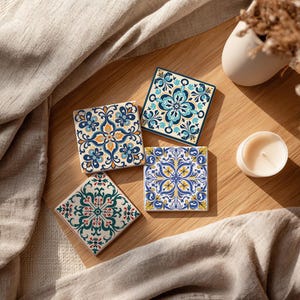 Op de afbeelding: Vier vierkante keramische onderzetters met ingewikkelde bloempatronen in blauw, groen, geel en oranje. De onderzetters liggen op een houten oppervlak, samen met een kleine kaars en een decoratieve vaas.
