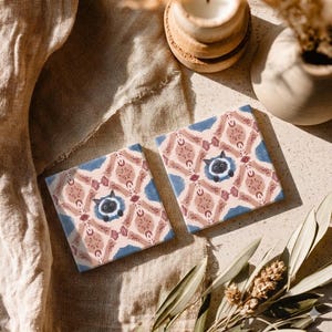 Può includere: Due sottobicchieri quadrati in ceramica con un motivo ripetuto di forme rosa, blu e marroni. Ogni sottobicchiere presenta un'illustrazione di un gatto nero al centro. I sottobicchieri sono esposti su una superficie dai toni neutri.