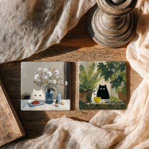 Op de afbeelding: Twee vierkante onderzetters met artistieke illustraties. De linker onderzetter toont een witte kat naast een blauwe vaas met witte bloemen en rode bessen. De rechter onderzetter toont een zwarte kat omringd door groene planten en een gele kom.