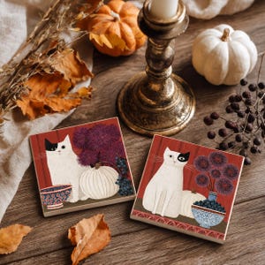 Könnte beinhalten: Zwei quadratische Untersetzer mit Illustrationen einer weißen Katze mit einem schwarzen Fleck über einem Auge, die neben einem weißen Kürbis, einer Schale mit dunklen Beeren und einer Vase mit violetten Blumen vor rotem Hintergrund stehen.