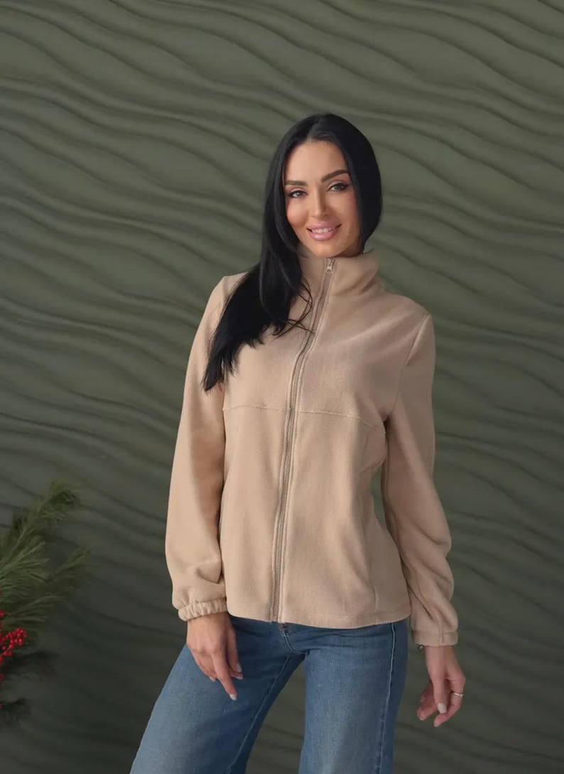 May include: A beige fleece jacket featuring a high collar, a full-length front zipper, and long sleeves with elasticized cuffs. The jacket is paired with blue denim jeans, creating a simple and functional outfit for cooler weather conditions.