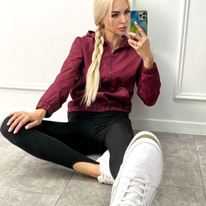 Puede incluir: Chaqueta con capucha y cremallera burdeos, leggings negros y zapatillas blancas. La chaqueta tiene una capucha con cordón y una cremallera completa. Los leggings son ajustados y las zapatillas son de perfil bajo con cordones blancos.