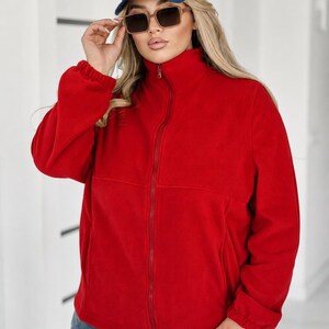 May include: A bright red fleece jacket with a high collar and a full-length front zipper, paired with a blue baseball cap featuring the white text ICON and brown-tinted sunglasses, worn with blue denim jeans with a distressed wash finish.