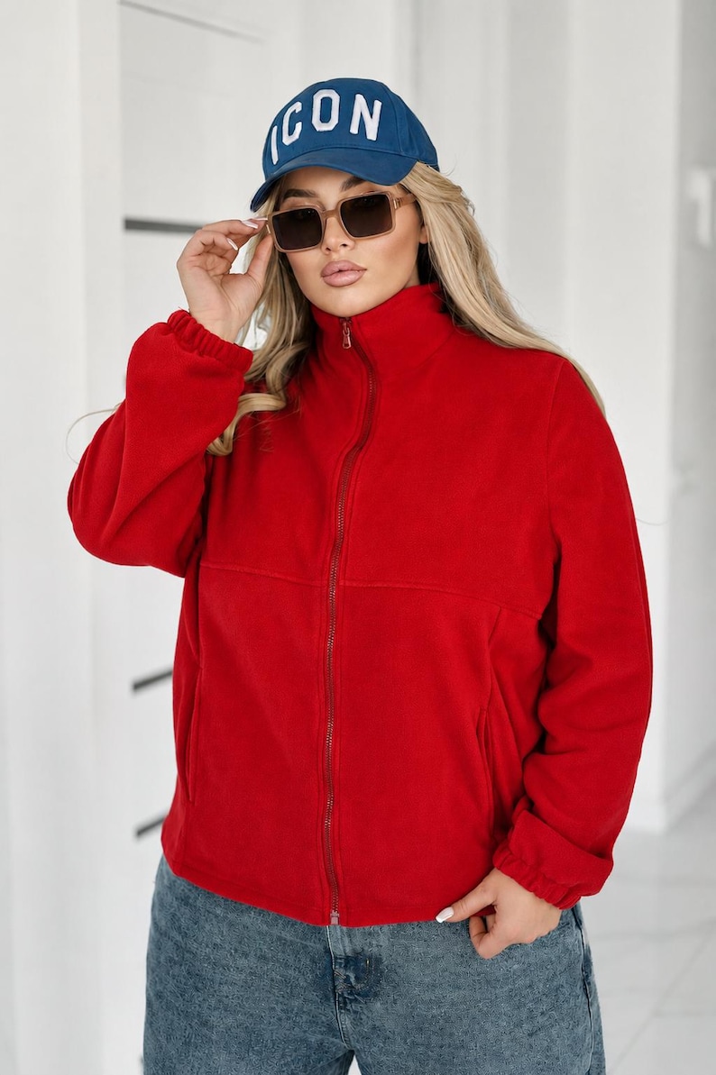 May include: A bright red fleece jacket with a high collar and a full-length front zipper, paired with a blue baseball cap featuring the white text ICON and brown-tinted sunglasses, worn with blue denim jeans with a distressed wash finish.