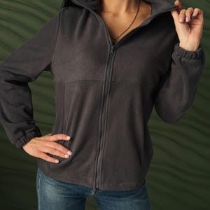May include: A dark charcoal gray fleece jacket featuring a full-length front zipper, a high collar, long sleeves with elasticized cuffs, and a horizontal seam detail across the chest. The jacket is paired with blue denim jeans.