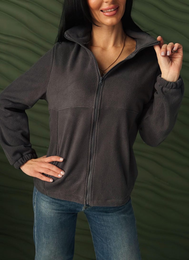 May include: A dark charcoal gray fleece jacket featuring a full-length front zipper, a high collar, long sleeves with elasticized cuffs, and a horizontal seam detail across the chest. The jacket is paired with blue denim jeans.