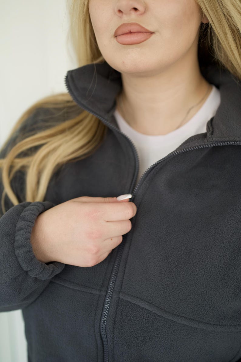 May include: A dark charcoal gray fleece jacket featuring a high collar, a full-length front zipper, and long sleeves with elasticized cuffs. The garment is worn over a plain white shirt, showcasing the soft texture of the fleece material.