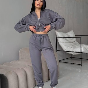 May include: A grey tracksuit, comprising a zip-up jacket and matching joggers. The jacket is cropped, and the joggers have a drawstring waist. The outfit is worn with white trainers. The set is made of a soft, comfortable material.