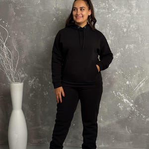 May include: A black hoodie and matching sweatpants set. The hoodie has a drawstring hood and a front pocket. The sweatpants have an elastic waistband and cuffs. The person is wearing white sneakers.