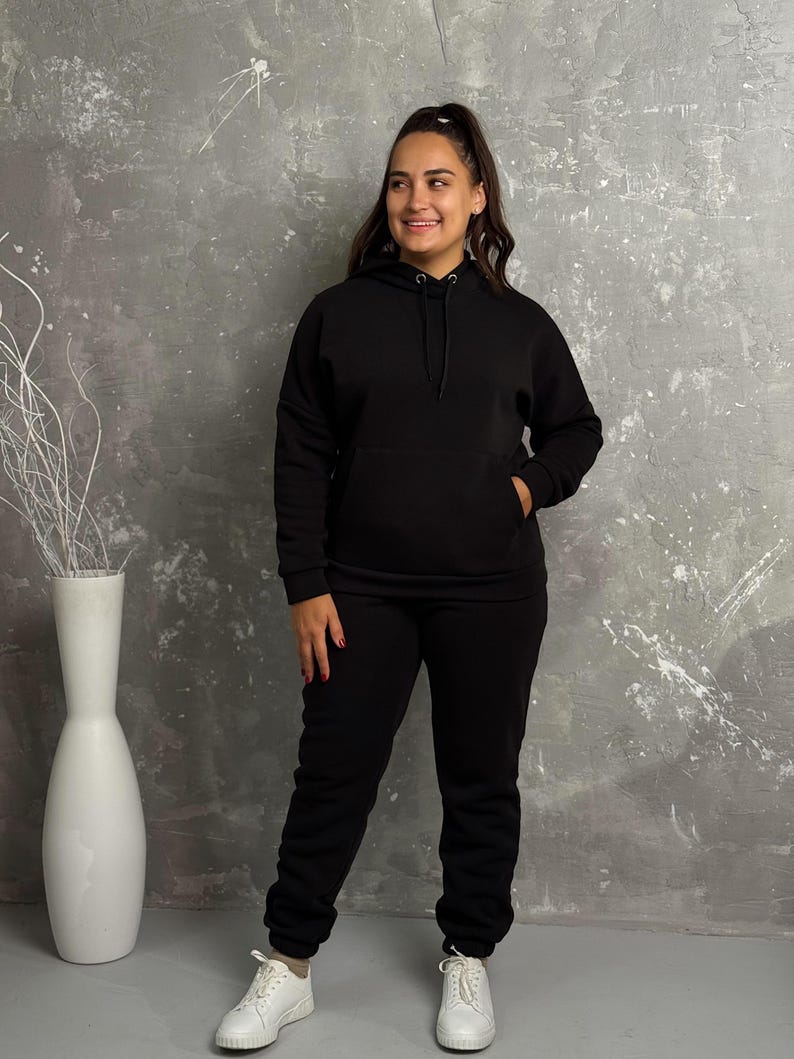 May include: A black hoodie and matching sweatpants set. The hoodie has a drawstring hood and a front pocket. The sweatpants have an elastic waistband and cuffs. The person is wearing white sneakers.