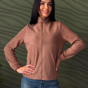 May include: A solid mocha brown fleece jacket featuring a high collar, a full-length front zipper, long sleeves with elasticized cuffs, and two side pockets. The jacket is worn over blue denim jeans, showcasing a simple and functional design.