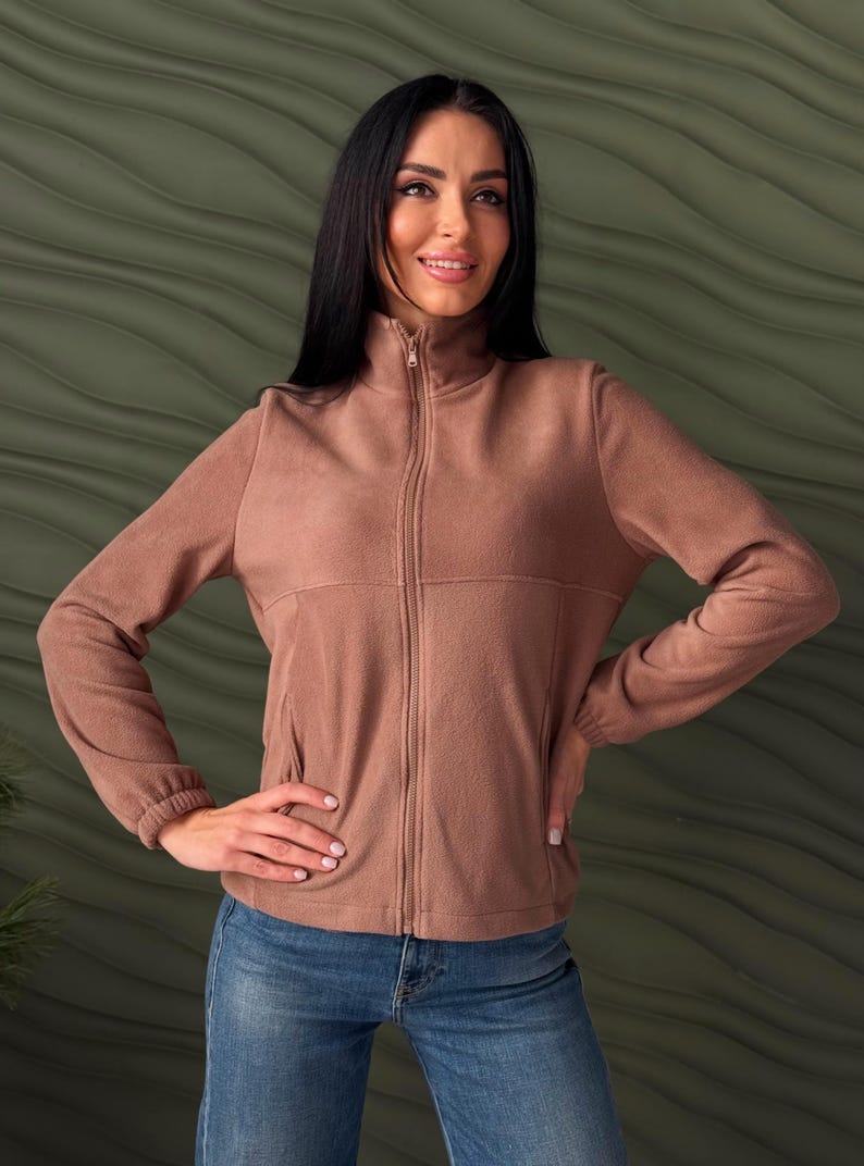 May include: A solid mocha brown fleece jacket featuring a high collar, a full-length front zipper, long sleeves with elasticized cuffs, and two side pockets. The jacket is worn over blue denim jeans, showcasing a simple and functional design.
