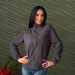May include: A dark gray fleece jacket featuring a full-length front zipper, a high mock neck collar, and long sleeves with elasticized cuffs. The jacket has a horizontal seam across the chest and is paired with blue denim jeans.