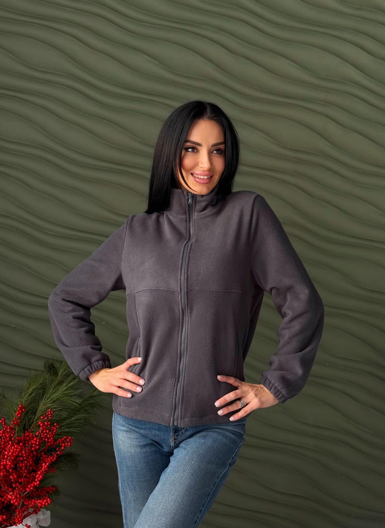 May include: A dark gray fleece jacket featuring a full-length front zipper, a high mock neck collar, and long sleeves with elasticized cuffs. The jacket has a horizontal seam across the chest and is paired with blue denim jeans.