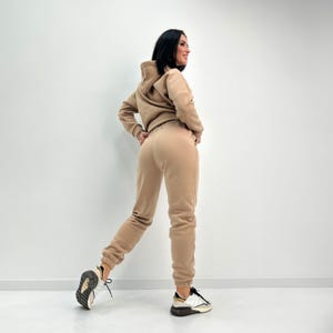 May include: A tan-colored hooded sweatshirt and matching sweatpants set. The outfit is made of a soft material and is worn with white and black sneakers. The model is standing in front of a white wall.