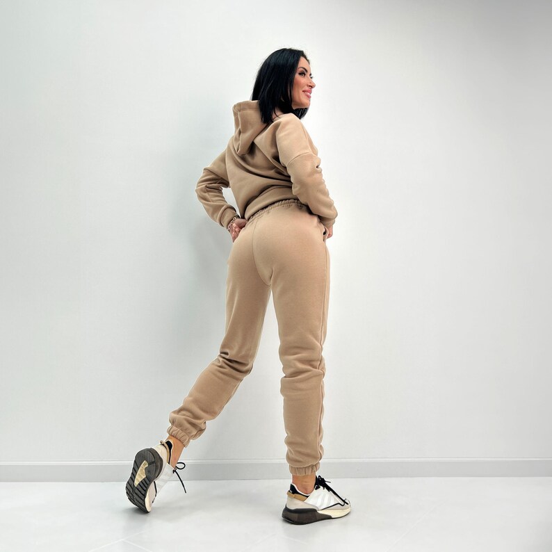May include: A tan-colored hooded sweatshirt and matching sweatpants set. The outfit is made of a soft material and is worn with white and black sneakers. The model is standing in front of a white wall.