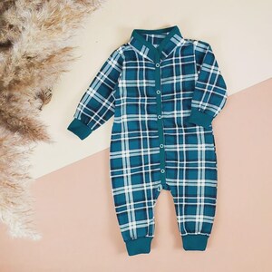 May include: A teal and white plaid baby onesie with long sleeves and a button-up front. The onesie has a collared neckline and teal cuffs and trim. The garment is displayed on a pink and beige background.