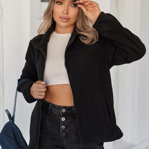 May include: A black fleece zip-up jacket with a high collar and side pockets, worn with a white cropped top, dark gray button-fly jeans, and a dark brown baseball cap featuring a repeating gold logo pattern and a prominent gold circular emblem.