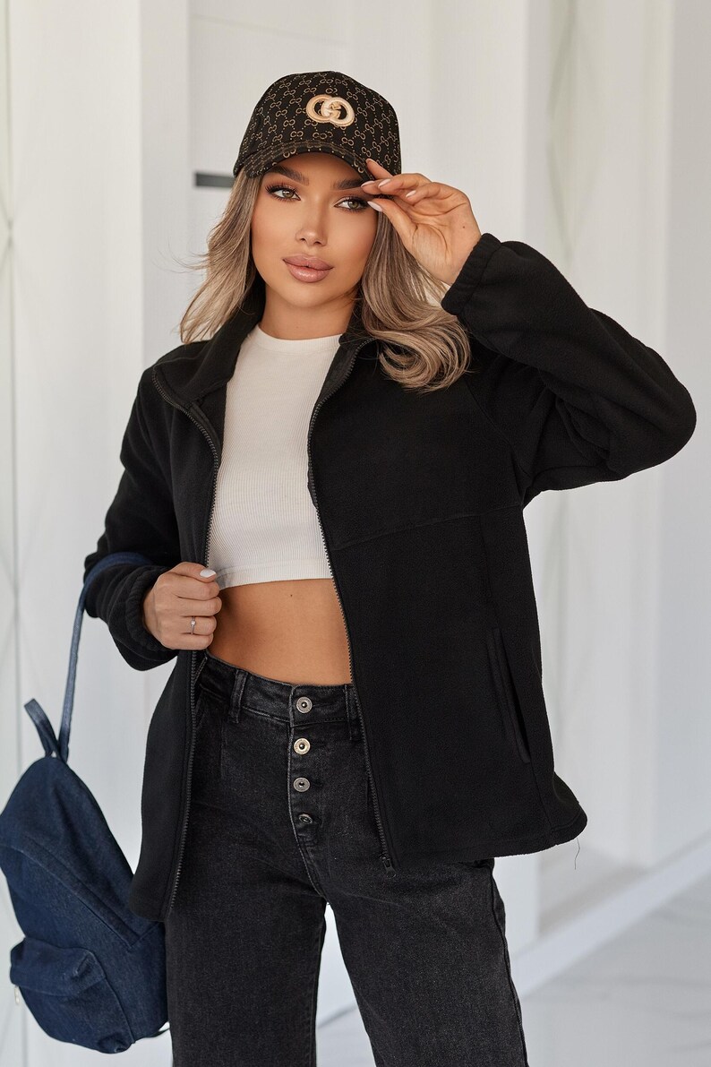 May include: A black fleece zip-up jacket with a high collar and side pockets, worn with a white cropped top, dark gray button-fly jeans, and a dark brown baseball cap featuring a repeating gold logo pattern and a prominent gold circular emblem.