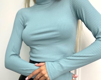 May include: A light blue ribbed turtleneck top with long sleeves. The top has a fitted silhouette and a high neckline. The fabric has vertical ribbing. The sleeves extend to the wrists. The top is paired with black bottoms.
