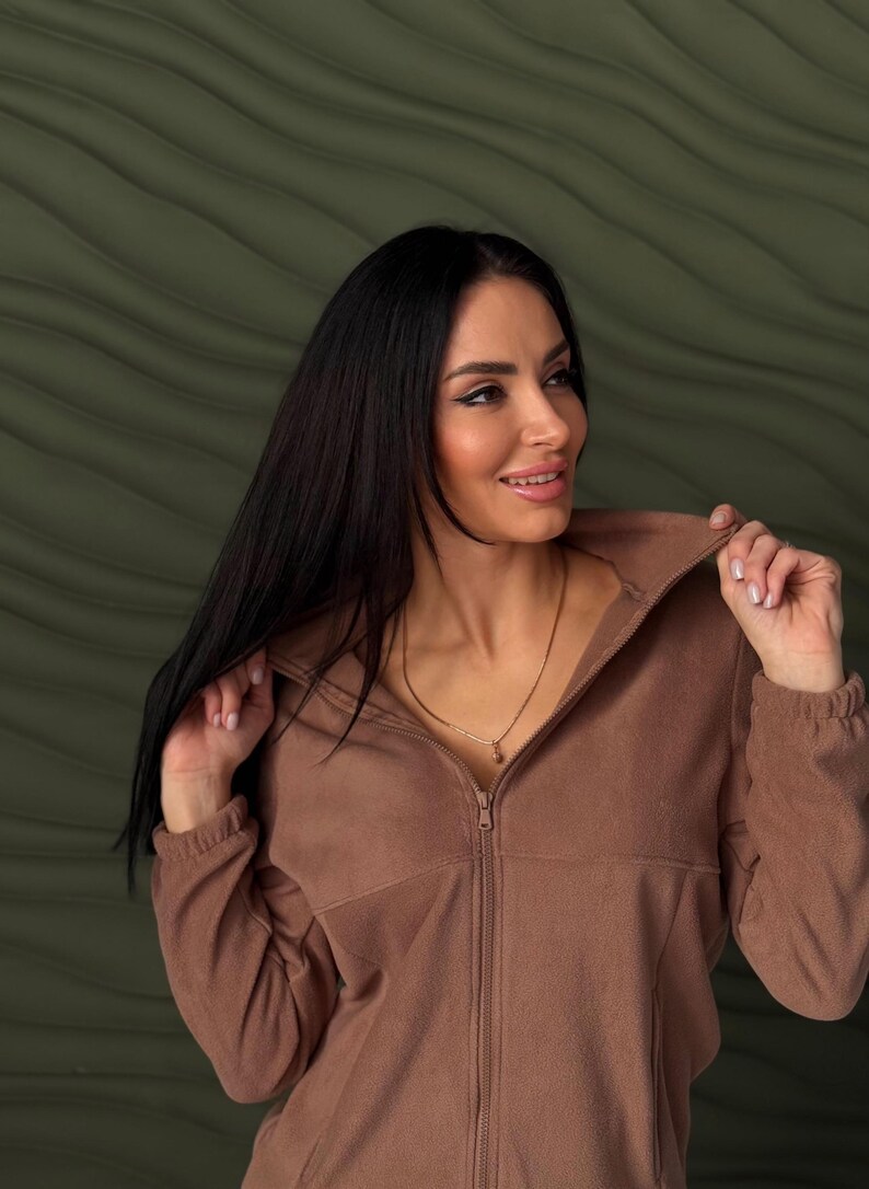 May include: A solid brown fleece jacket featuring a high collar, a full-length front zipper closure, and long sleeves with elasticized cuffs. The garment is constructed from a soft, textured fleece material with a consistent matte finish.