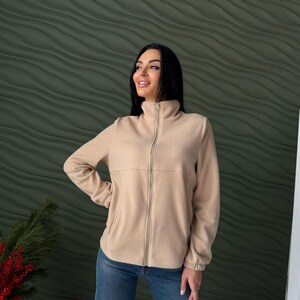 May include: A beige fleece jacket featuring a high collar, a full-length front zipper, long sleeves with elasticized cuffs, and two side pockets, paired with blue denim jeans.