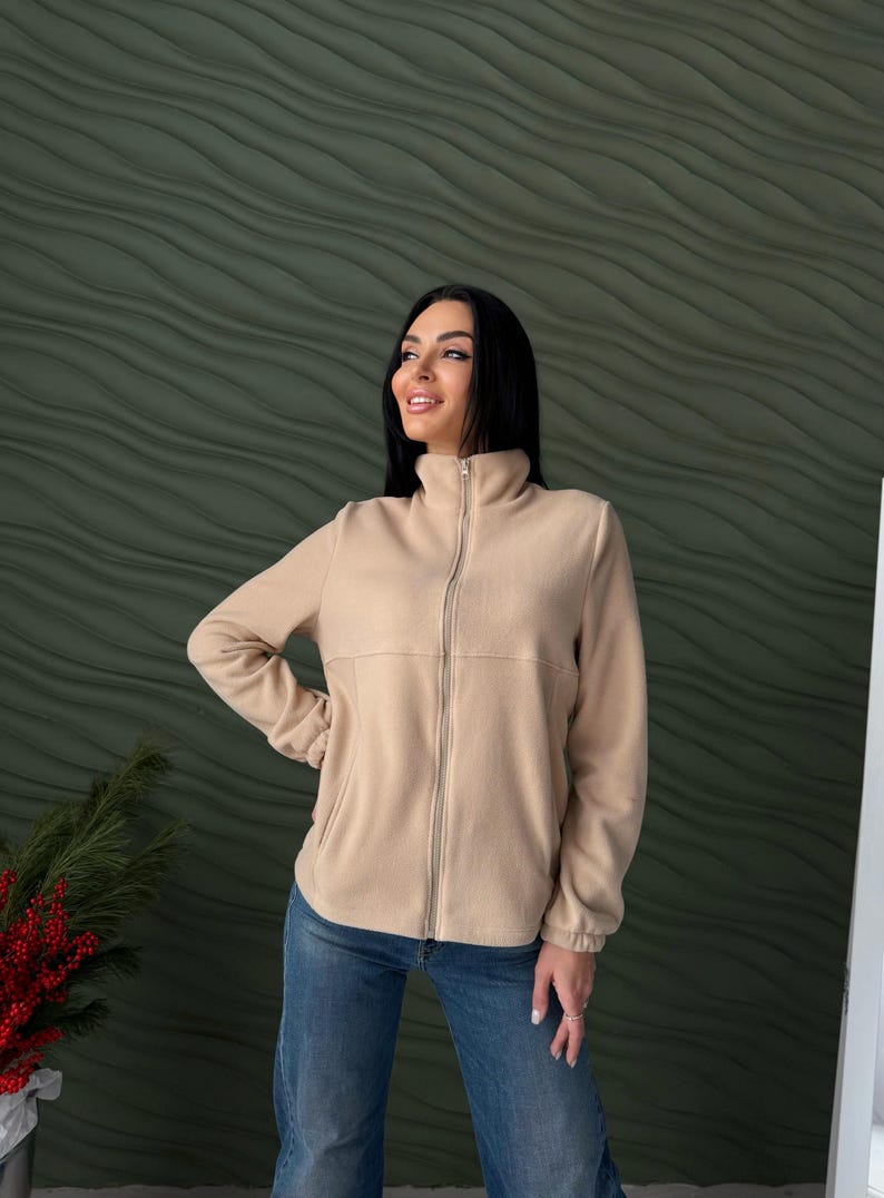 May include: A beige fleece jacket featuring a high collar, a full-length front zipper, long sleeves with elasticized cuffs, and two side pockets, paired with blue denim jeans.