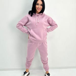 May include: A light purple hoodie and matching sweatpants set. The hoodie has a front pocket and a drawstring. The sweatpants are gathered at the ankles. The person is wearing white and black sneakers.