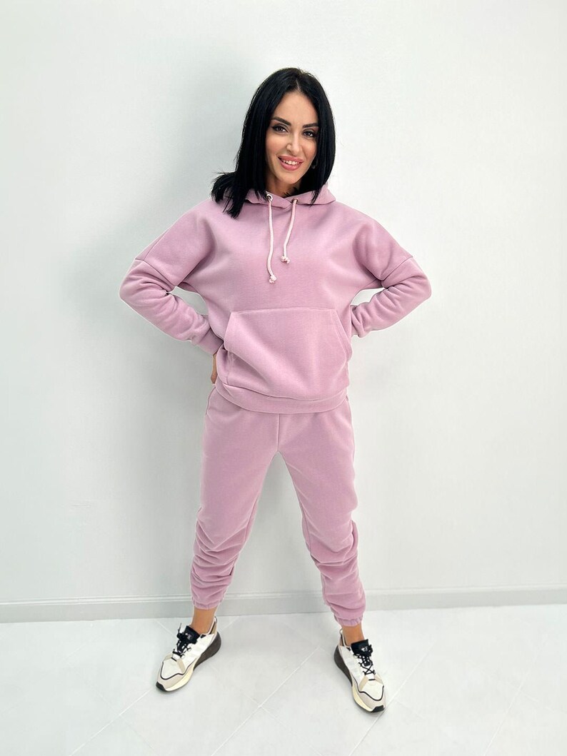 May include: A light purple hoodie and matching sweatpants set. The hoodie has a front pocket and a drawstring. The sweatpants are gathered at the ankles. The person is wearing white and black sneakers.